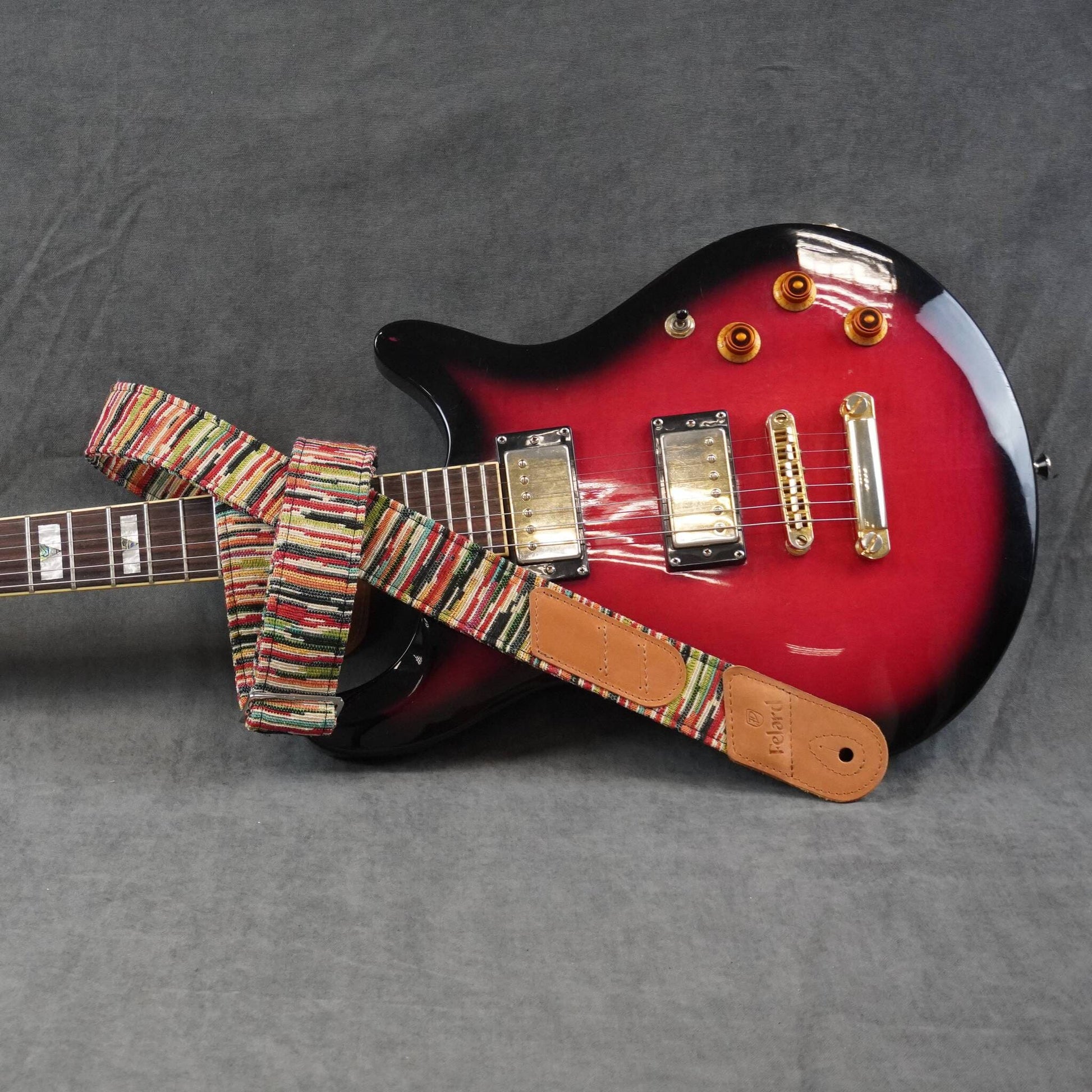 Colorful striped guitar strap featuring a tan leather patch, photographed on a gray textured fabric background.