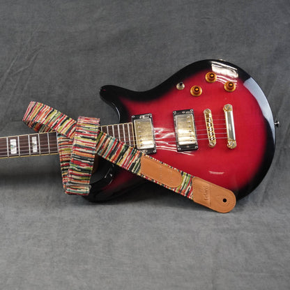 Colorful striped guitar strap featuring a tan leather patch, photographed on a gray textured fabric background.