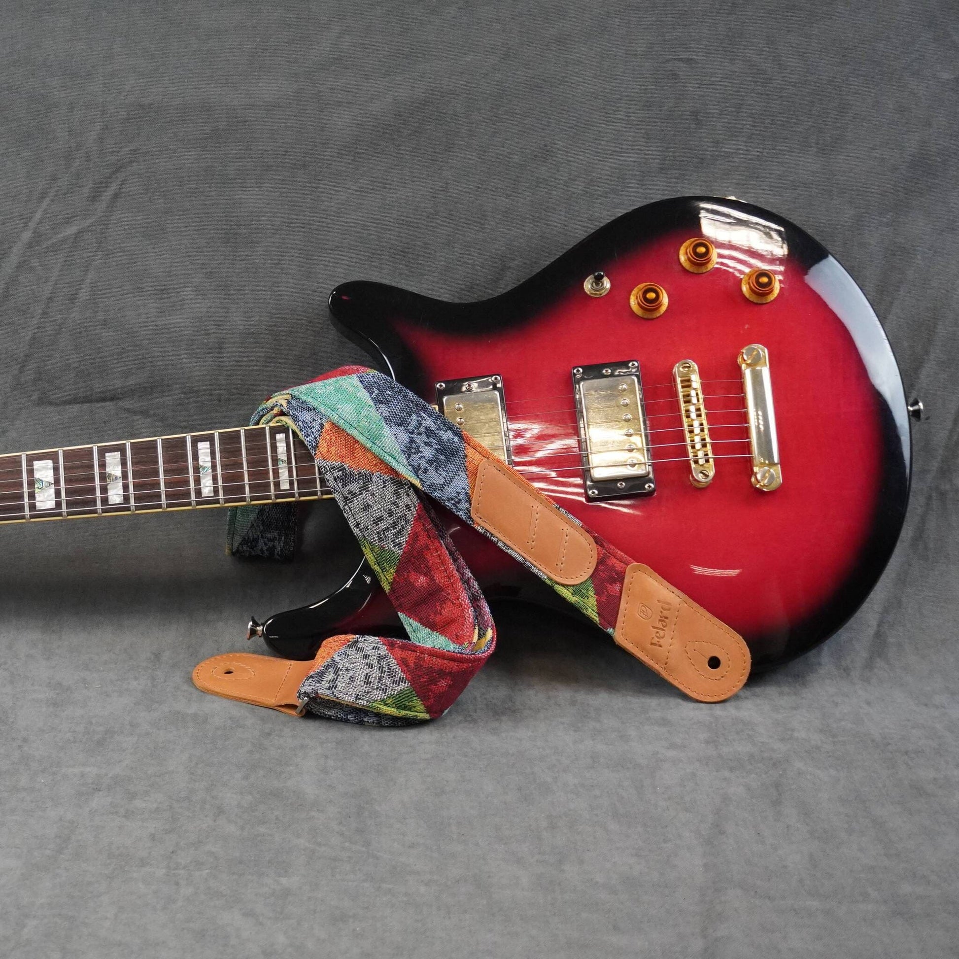 colorful patterned guitar strap featuring tan leather ends, displayed on a gray fabric background.