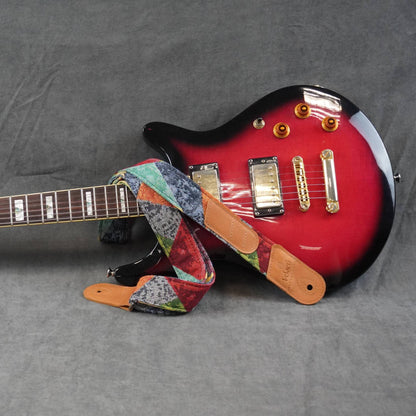 colorful patterned guitar strap featuring tan leather ends, displayed on a gray fabric background.