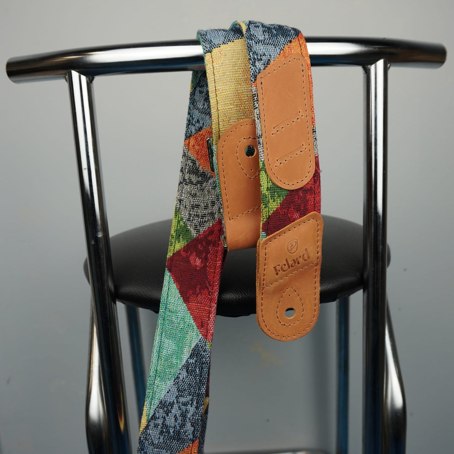 A colorful patchwork-patterned camera strap with tan leather accents hanging from a black chair against a gray background. The strap features the 'Focalland' brand name embossed on the leather tabs.