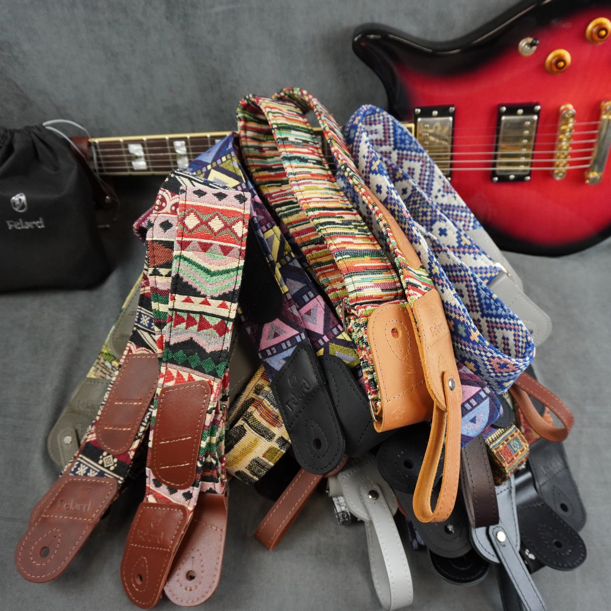 A pile of colorful guitar straps with geometric and striped patterns, brown leather ends, and metal adjusters, arranged on a gray surface with a red electric guitar and a black pouch in the background.