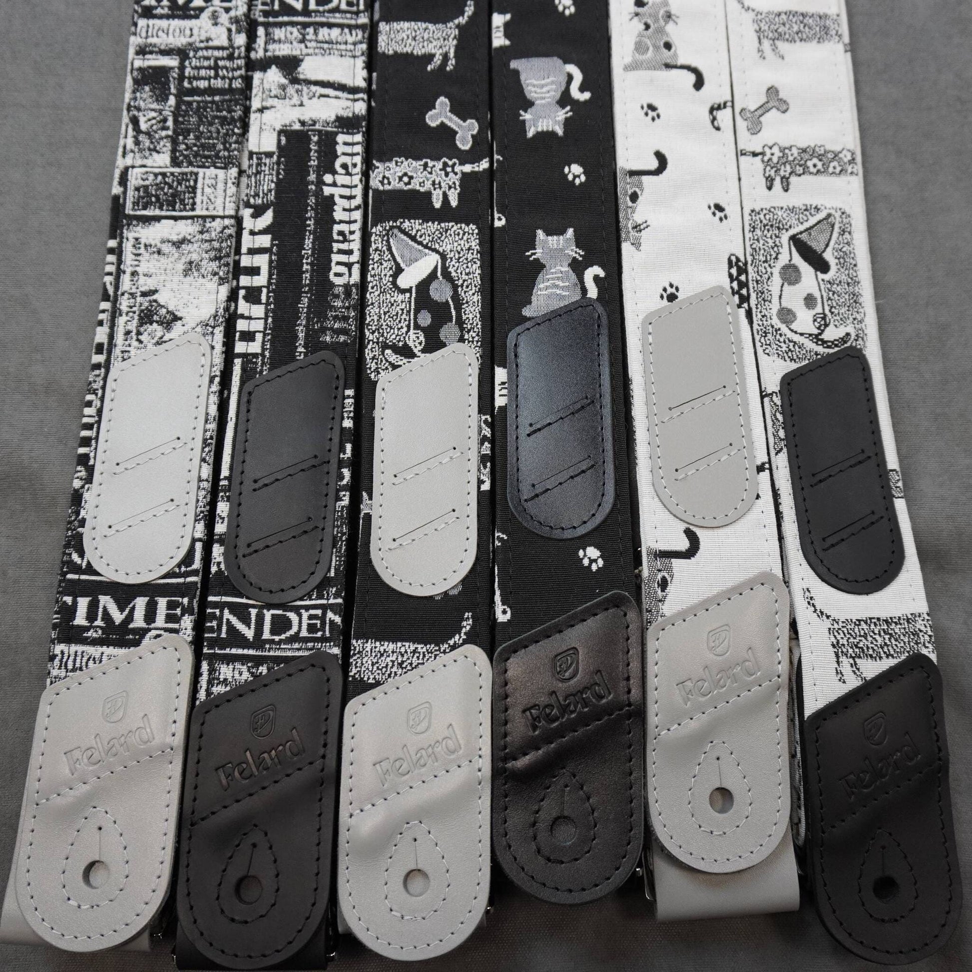Guitar straps laid flat on a gray surface, featuring black and white patterns including newspaper clippings, paw prints, cat silhouettes, and dog illustrations, with black and white leather end caps.