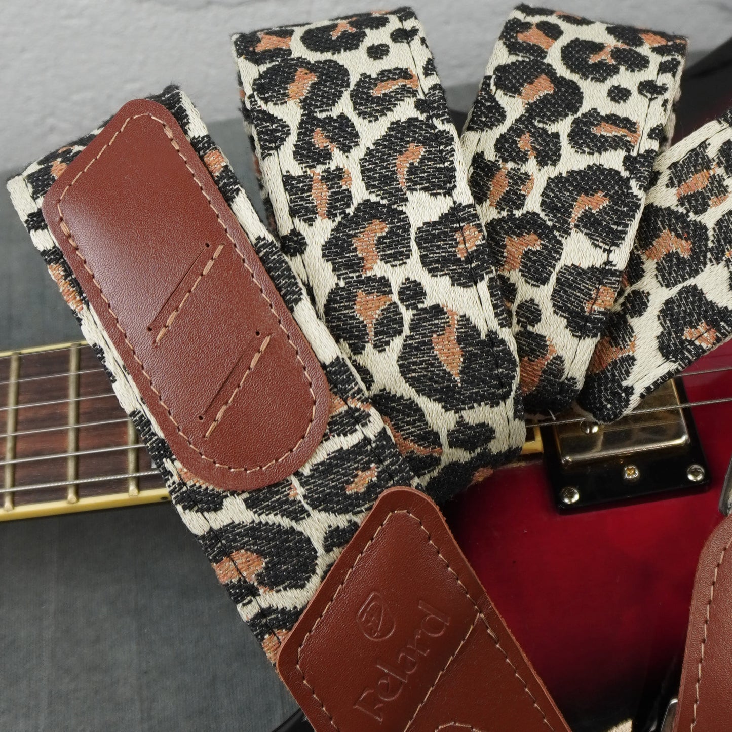 Close-up of a guitar strap with a leopard print pattern in beige, black, and brown, featuring brown faux-leather end pieces. The strap is shown resting on a red guitar body, with part of the guitar neck visible.