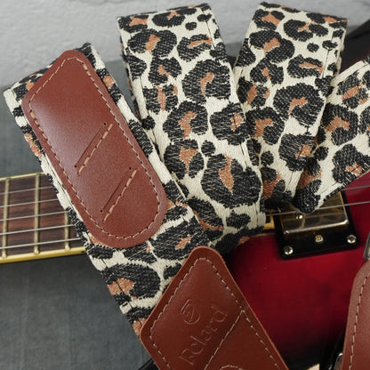 Close-up of a guitar strap with a leopard print pattern in beige, black, and brown, featuring brown faux-leather end pieces. The strap is shown resting on a red guitar body, with part of the guitar neck visible.