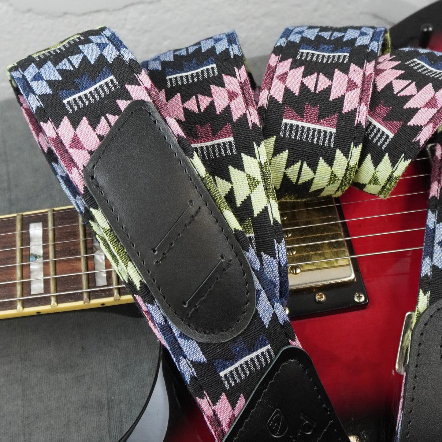 guitar strap with a colorful geometric pattern in pink, blue, green, and white on a black base, featuring black leather patches, shown draped over a red electric guitar.