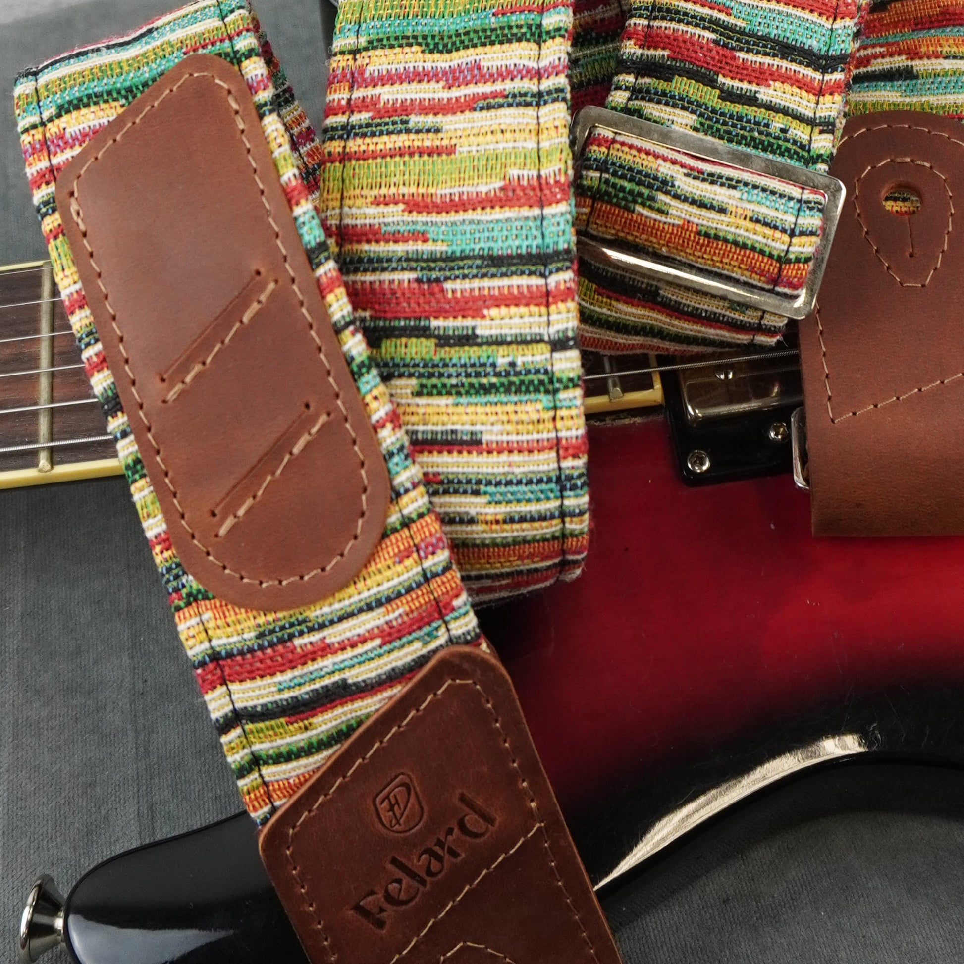 A close-up of a guitar strap with a colorful striped woven fabric and brown leather ends, shown draped over a red electric guitar. The brand name 'Felard' is visible on the leather end piece.