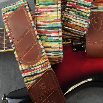 A close-up of a guitar strap with a colorful striped woven fabric and brown leather ends, shown draped over a red electric guitar. The brand name 'Felard' is visible on the leather end piece.