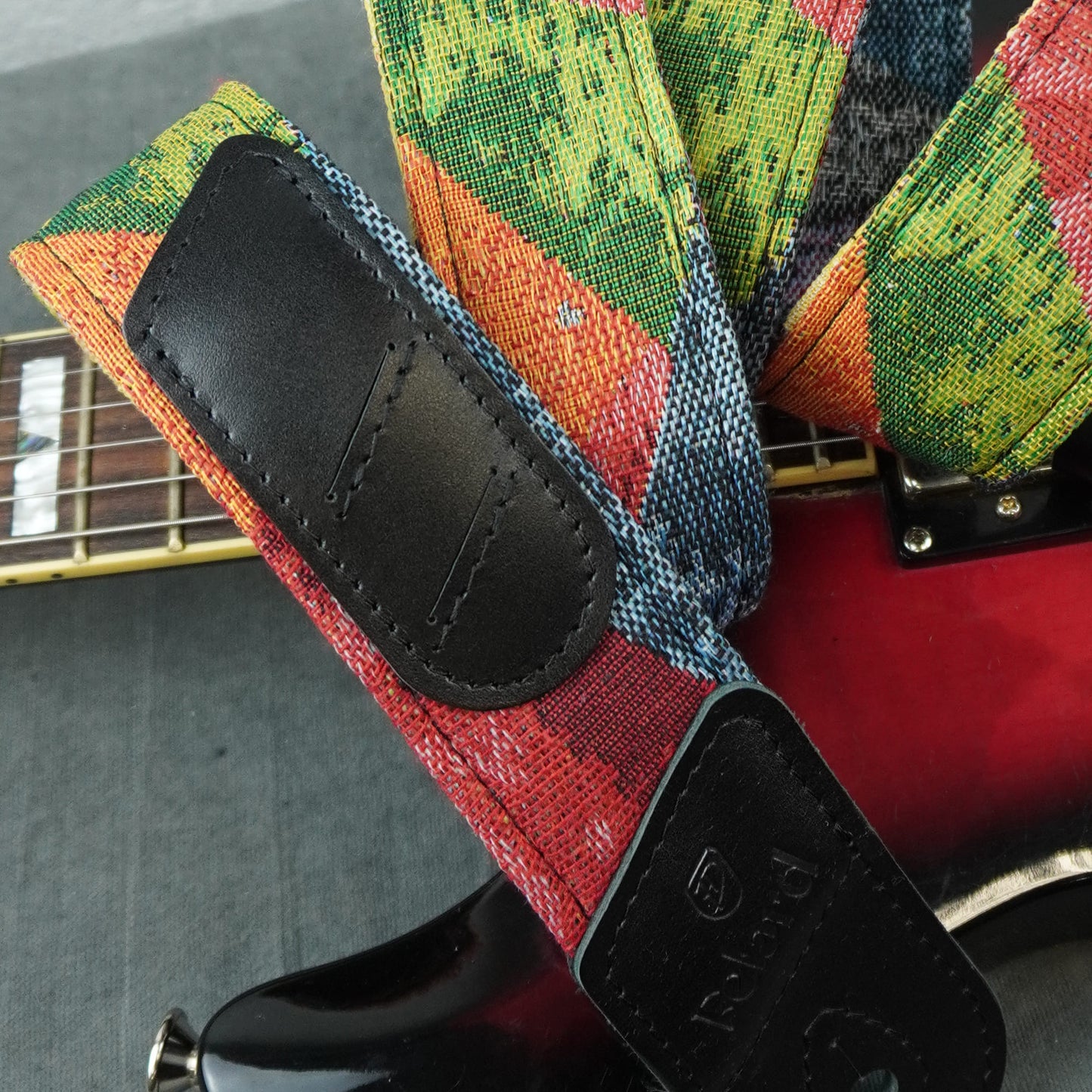 A close-up of a guitar strap with a colorful woven pattern in red, green, blue, and orange, featuring black leather patches. The strap is shown attached to a red electric guitar, with the guitar's neck and body visible against a gray background.