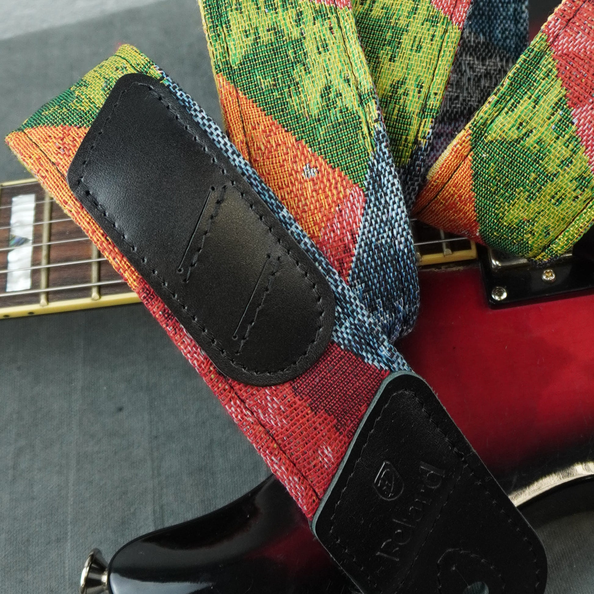 A close-up of a guitar strap with a colorful woven pattern in red, green, blue, and orange, featuring black leather patches. The strap is shown attached to a red electric guitar, with the guitar's neck and body visible against a gray background.