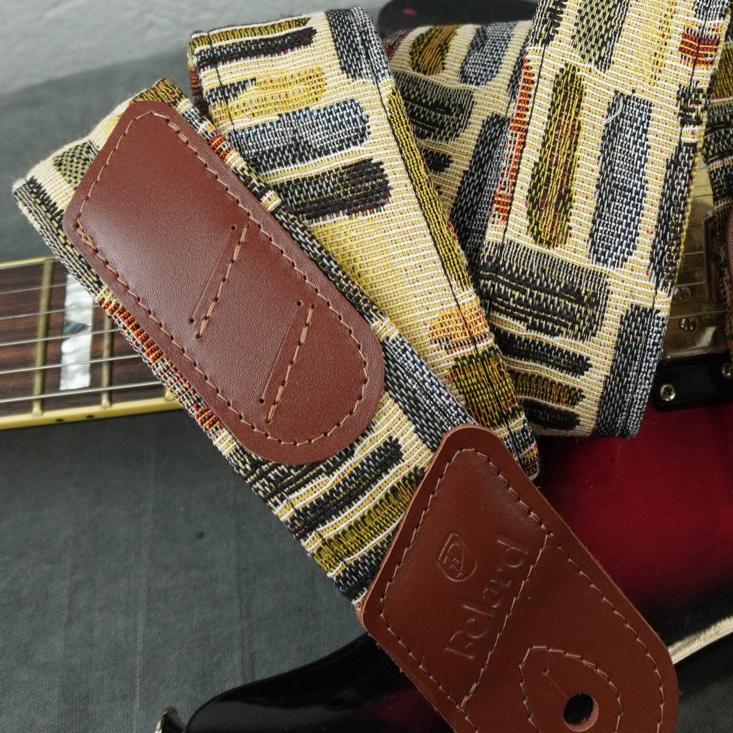 A close-up of a guitar strap with a woven geometric pattern in beige, black, yellow, and gray, featuring brown leather patches. The strap is draped over a guitar body.