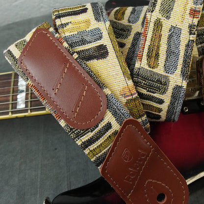 A close-up of a guitar strap with a woven geometric pattern in beige, black, yellow, and gray, featuring brown leather patches. The strap is draped over a guitar body.