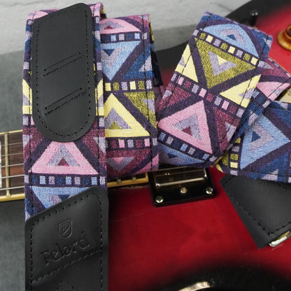 A close-up of a guitar strap with a colorful geometric pattern in pink, blue, yellow, and purple, featuring black faux-leather end pieces and a metal clamp, shown resting on a red electric guitar.