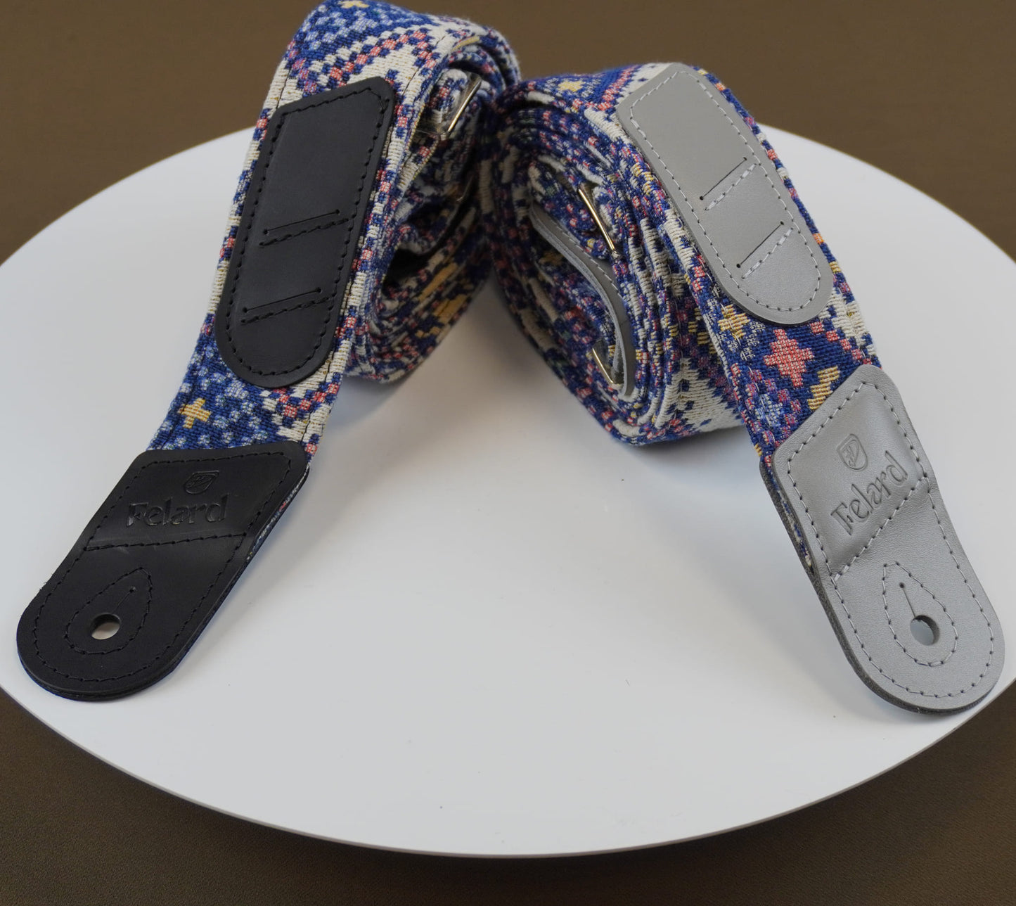 Guitar strap with a blue, purple, and red geometric pattern, featuring black leather end caps and a light gray adjustable buckle, displayed on a white circular plate.