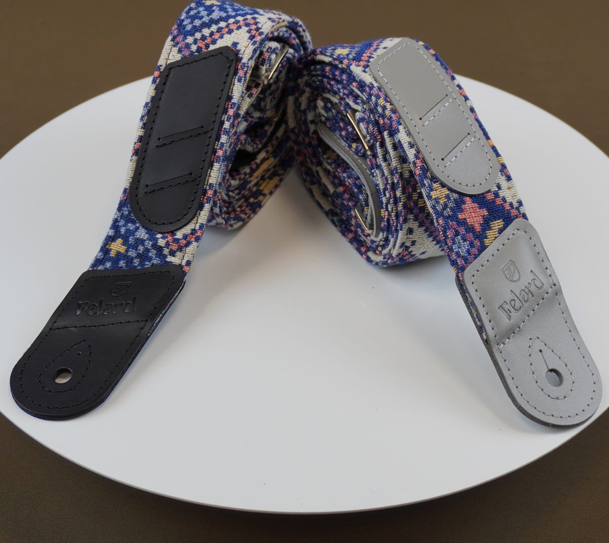 Guitar strap with a blue, purple, and red geometric pattern, featuring black leather end caps and a light gray adjustable buckle, displayed on a white circular plate.