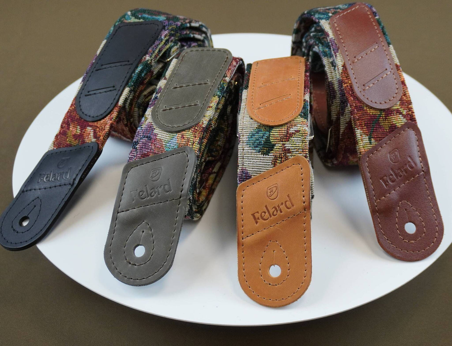 Four Felard guitar strap fabric samples with different leather end caps are arranged on a white plate against a dark brown background.