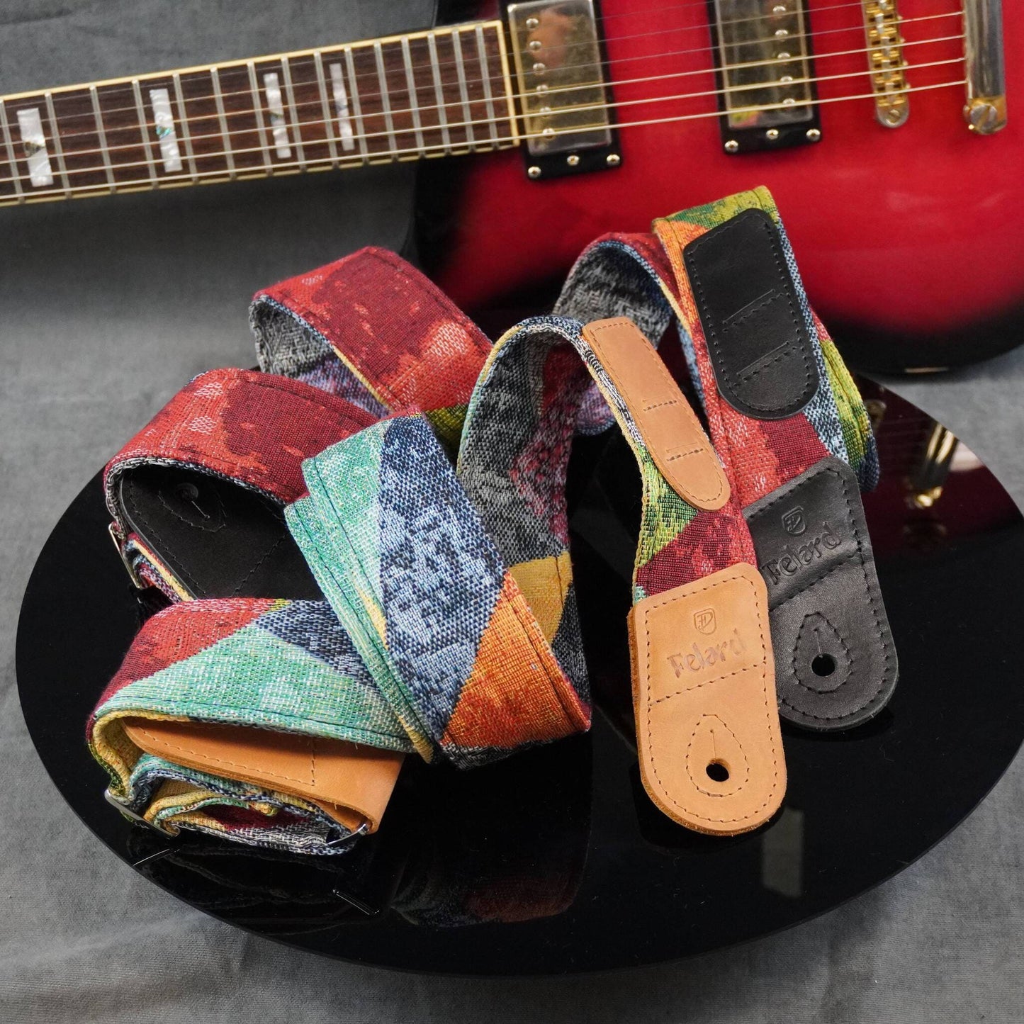 A guitar strap with a multicolor patchwork pattern and tan leather end pieces, displayed on a black reflective surface with a red electric guitar in the background.