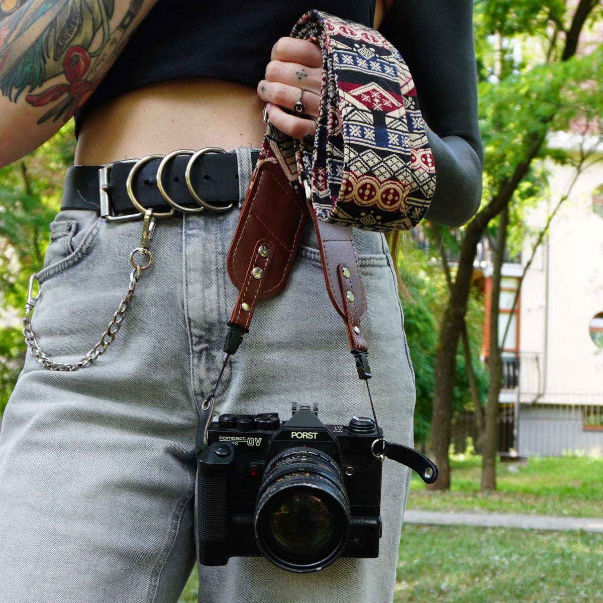 A person outdoors holding a brown leather camera strap with a patterned pouch. A black PORST camera hangs from the strap