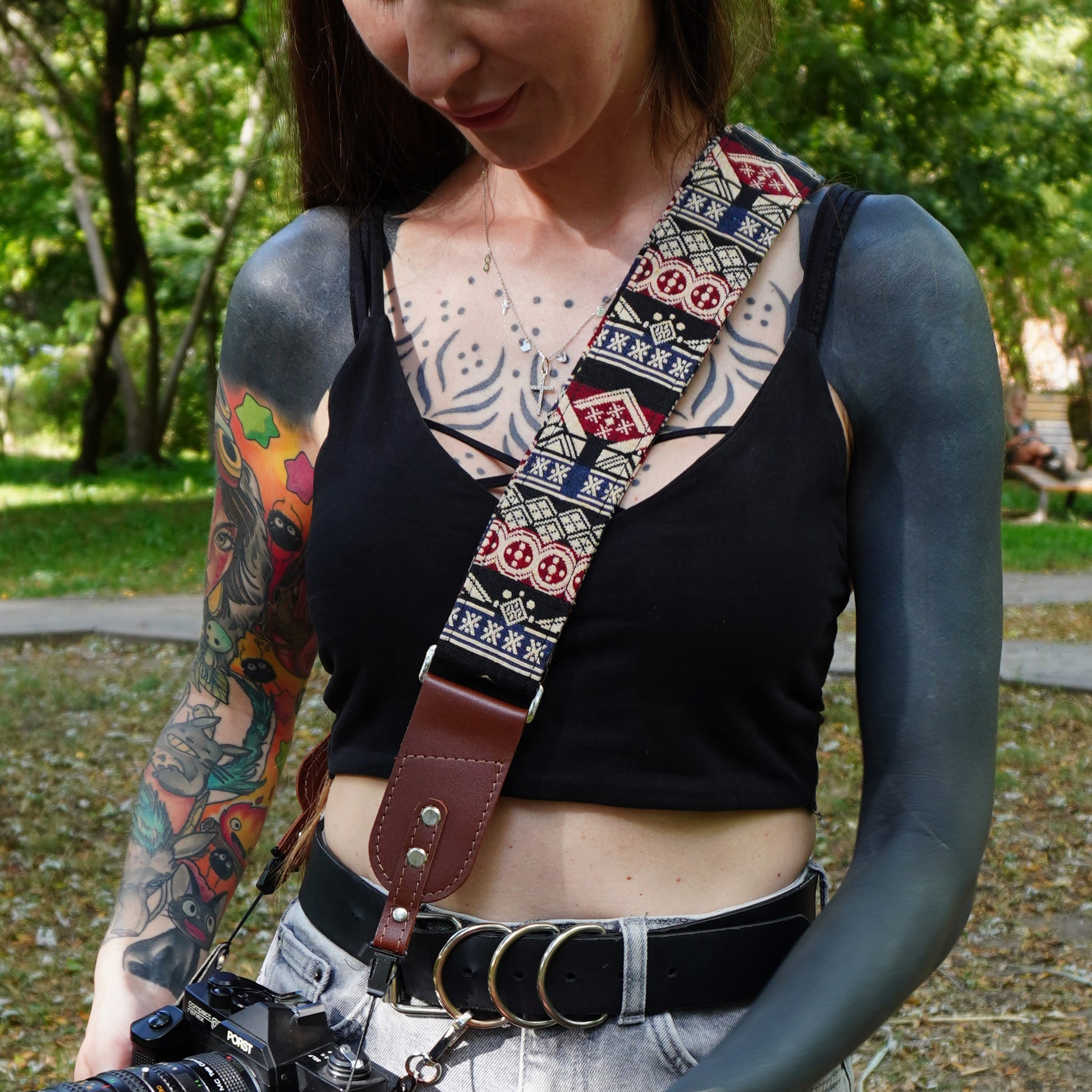 A woman outdoors wearing a patterned camera strap with a brown leather loop, carrying a camera over her shoulder. The strap features a geometric tribal-style pattern in red, blue, white, and black.