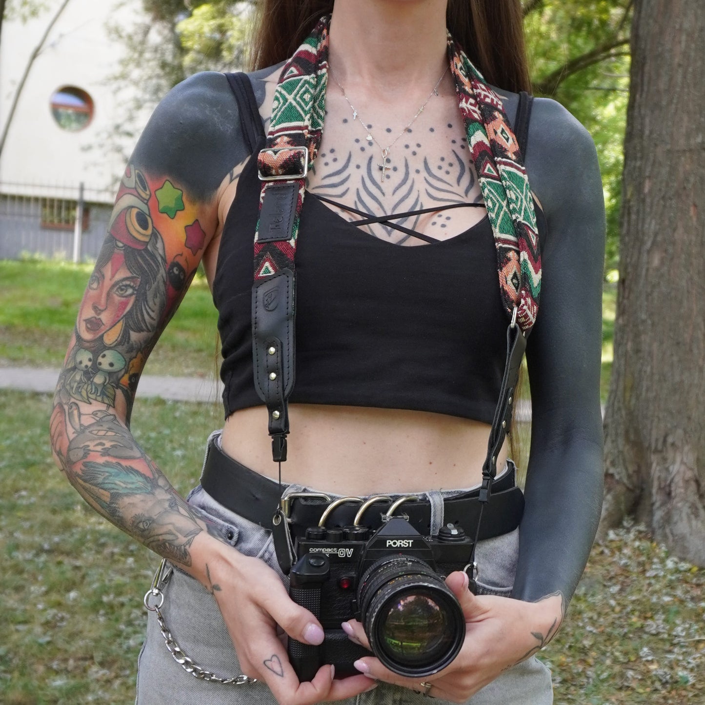 A person outdoors holding a vintage camera with a patterned fabric camera strap draped over their shoulders.