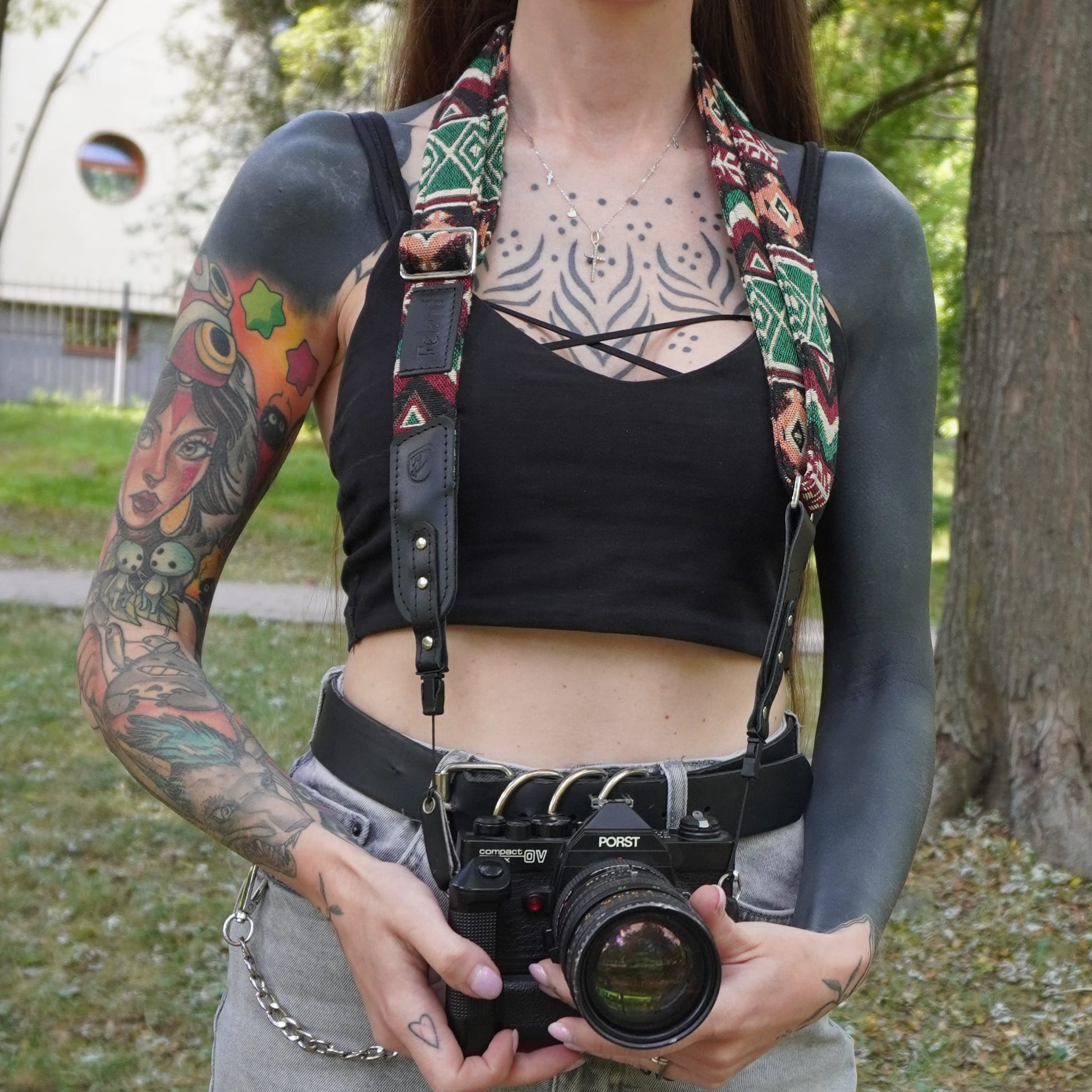 A person outdoors holding a vintage camera with a patterned fabric camera strap draped over their shoulders.