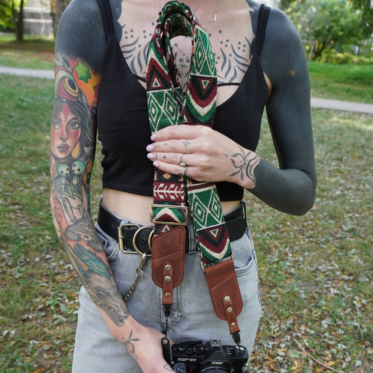 A tattooed person outdoors wearing a patterned camera strap with brown leather ends and silver hardware, holding a camera. The strap has a Southwestern geometric design in green, red, and cream.