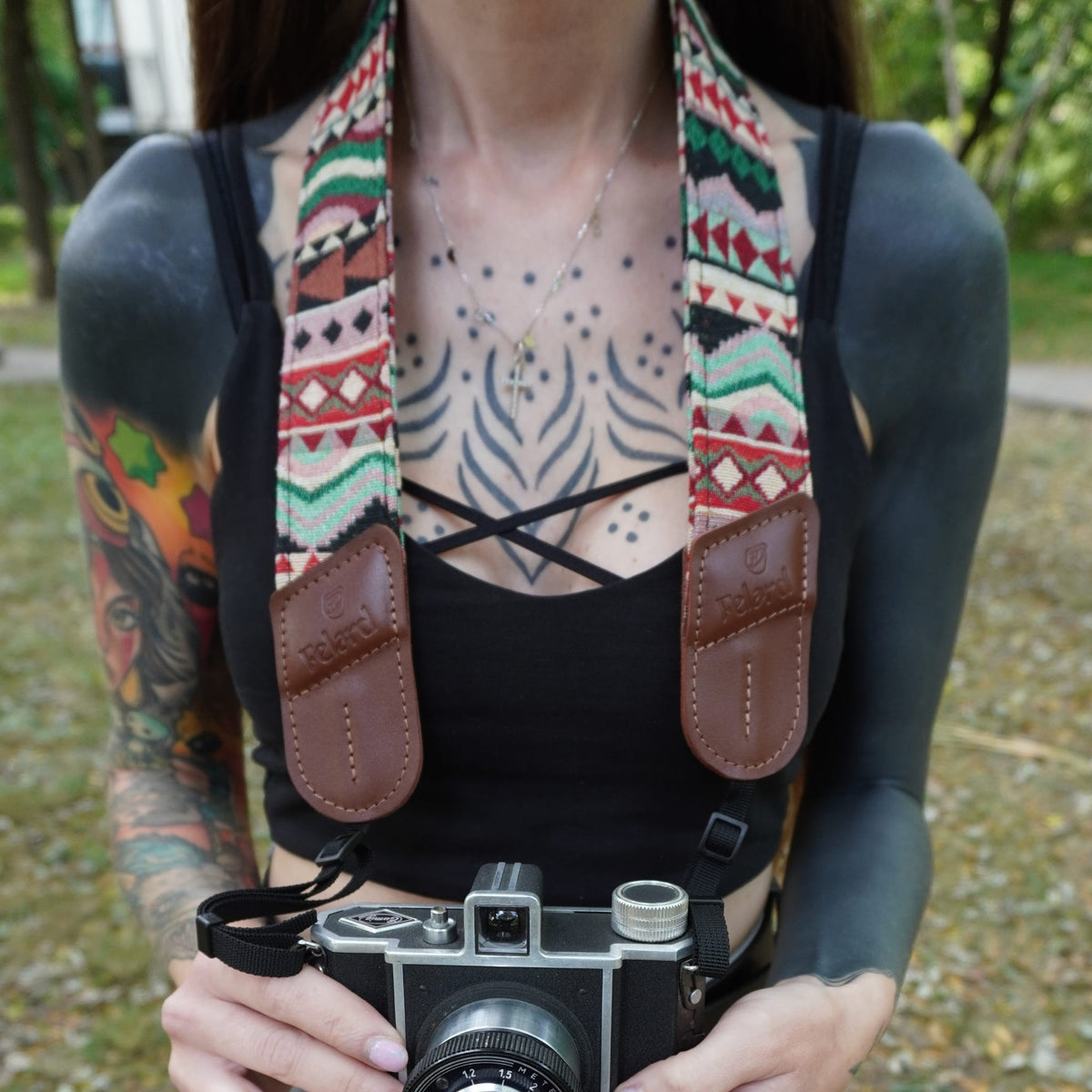 A close-up lifestyle shot of a person wearing a colorful geometric patterned camera strap with brown leather attachments, holding a vintage camera outdoors.