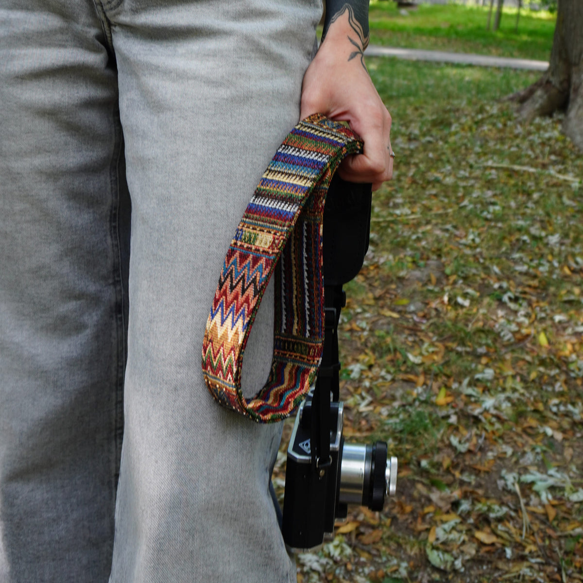 A woman outdoors wearing a patterned camera strap with a leather details, carrying a camera over her shoulder. The strap features a geometric boho-style pattern.