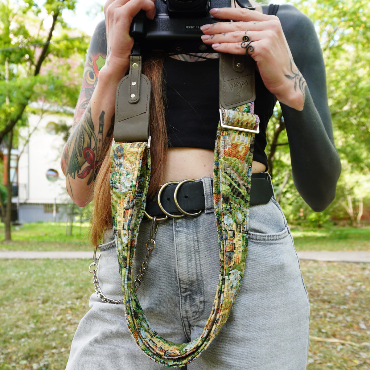 A person outdoors holding a camera with a wide, colorful patterned camera strap draped around the waist. The strap features a multicolor floral and abstract print in green, yellow, orange, and blue, with olive green leather accents and a silver chain link detail.