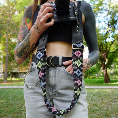 A close-up lifestyle shot of a person outdoors wearing a black leather camera strap with a colorful geometric patterned webbing section. The person is holding a DSLR camera in front of their face with trees in the background.