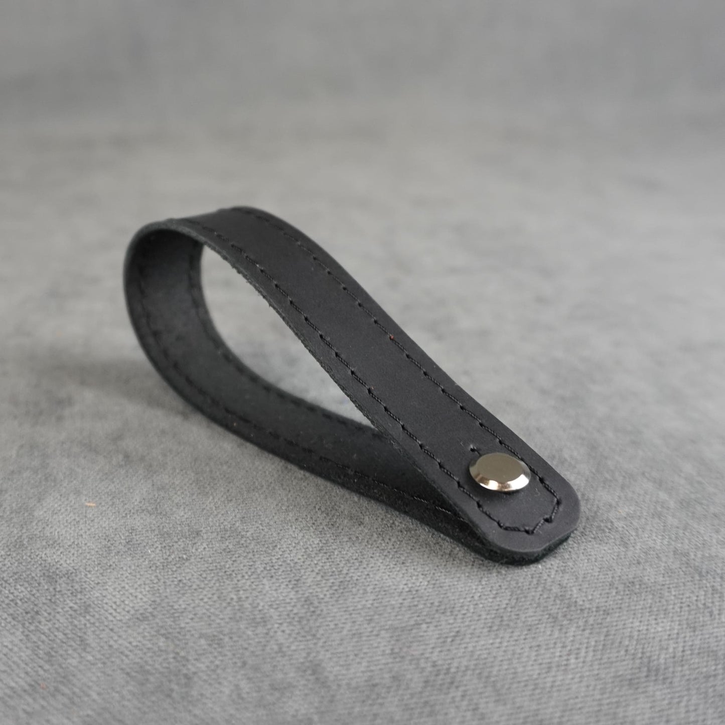 A black matte leather strap headstock adapter with a single silver metal rivet, photographed on a textured gray background.