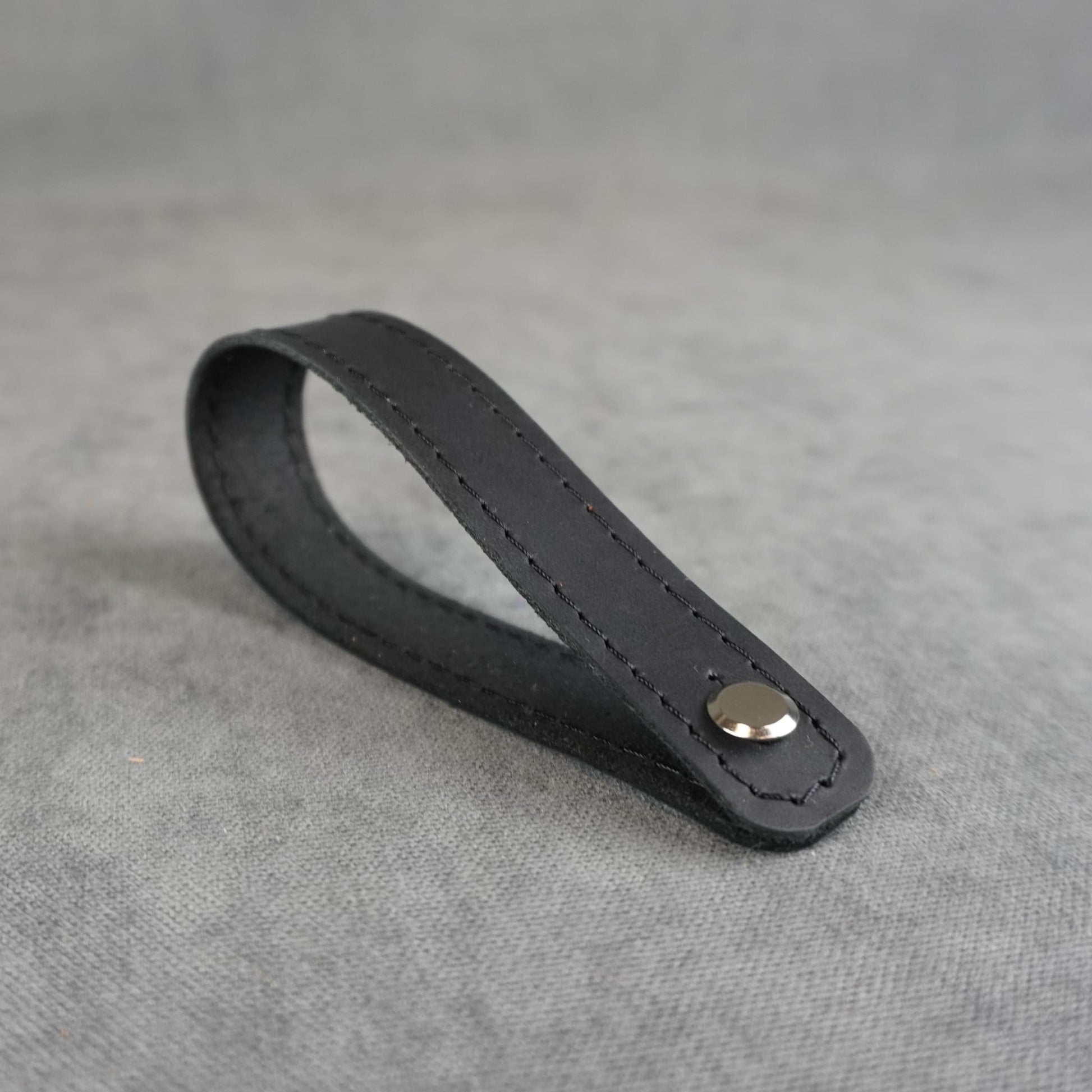 A black matte leather strap headstock adapter with a single silver metal rivet, photographed on a textured gray background.