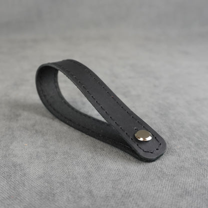 A black matte leather strap headstock adapter with a single silver metal rivet, photographed on a textured gray background.