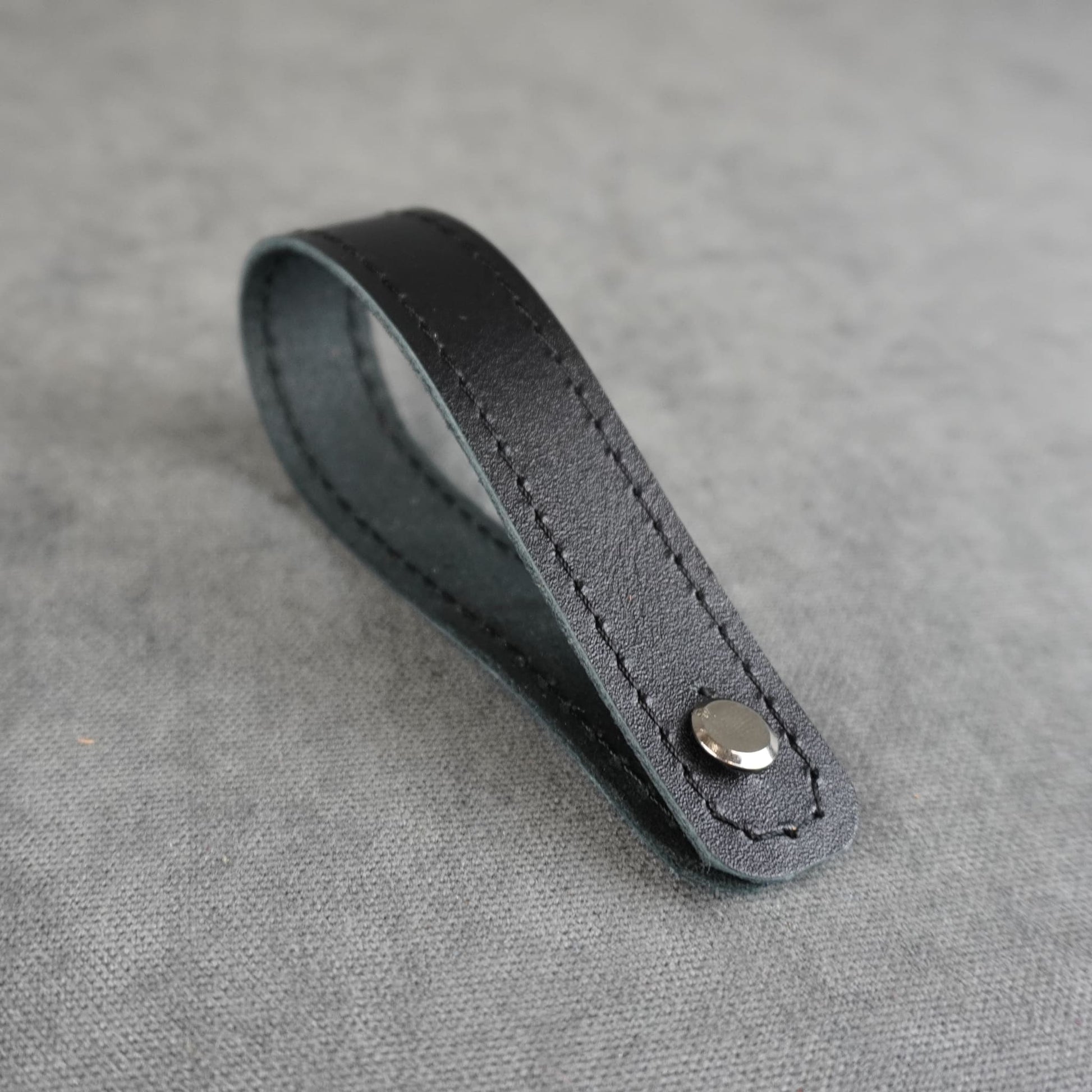 A black smooth leather strap headstock adapter with a single silver metal rivet, photographed on a textured gray background.