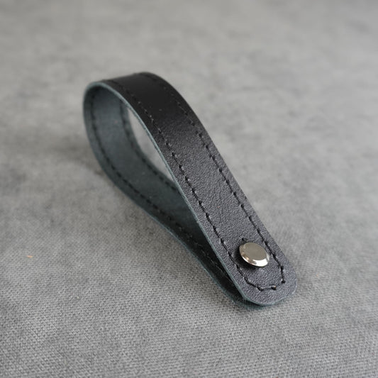 A black smooth leather strap headstock adapter with a single silver metal rivet, photographed on a textured gray background.