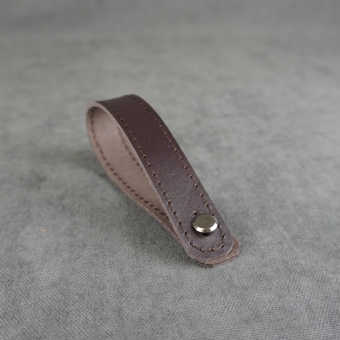 A dark brown smooth leather strap headstock adapter with a single silver metal rivet, photographed on a textured gray background