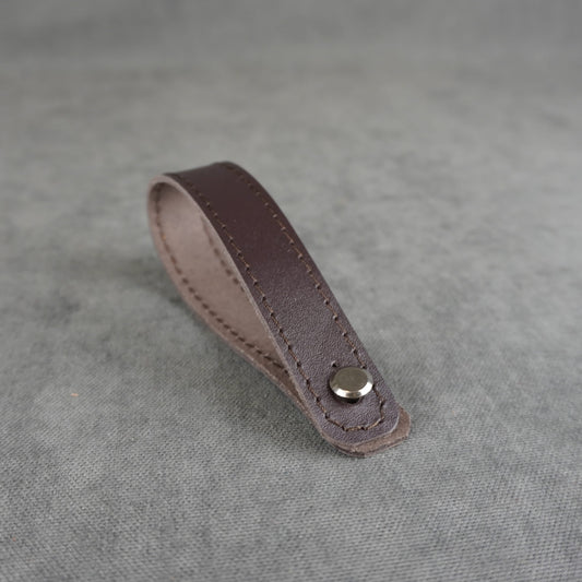 A dark brown smooth leather strap headstock adapter with a single silver metal rivet, photographed on a textured gray background