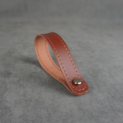 A cognac smooth leather strap headstock adapter with a single silver metal rivet, photographed on a textured gray background.