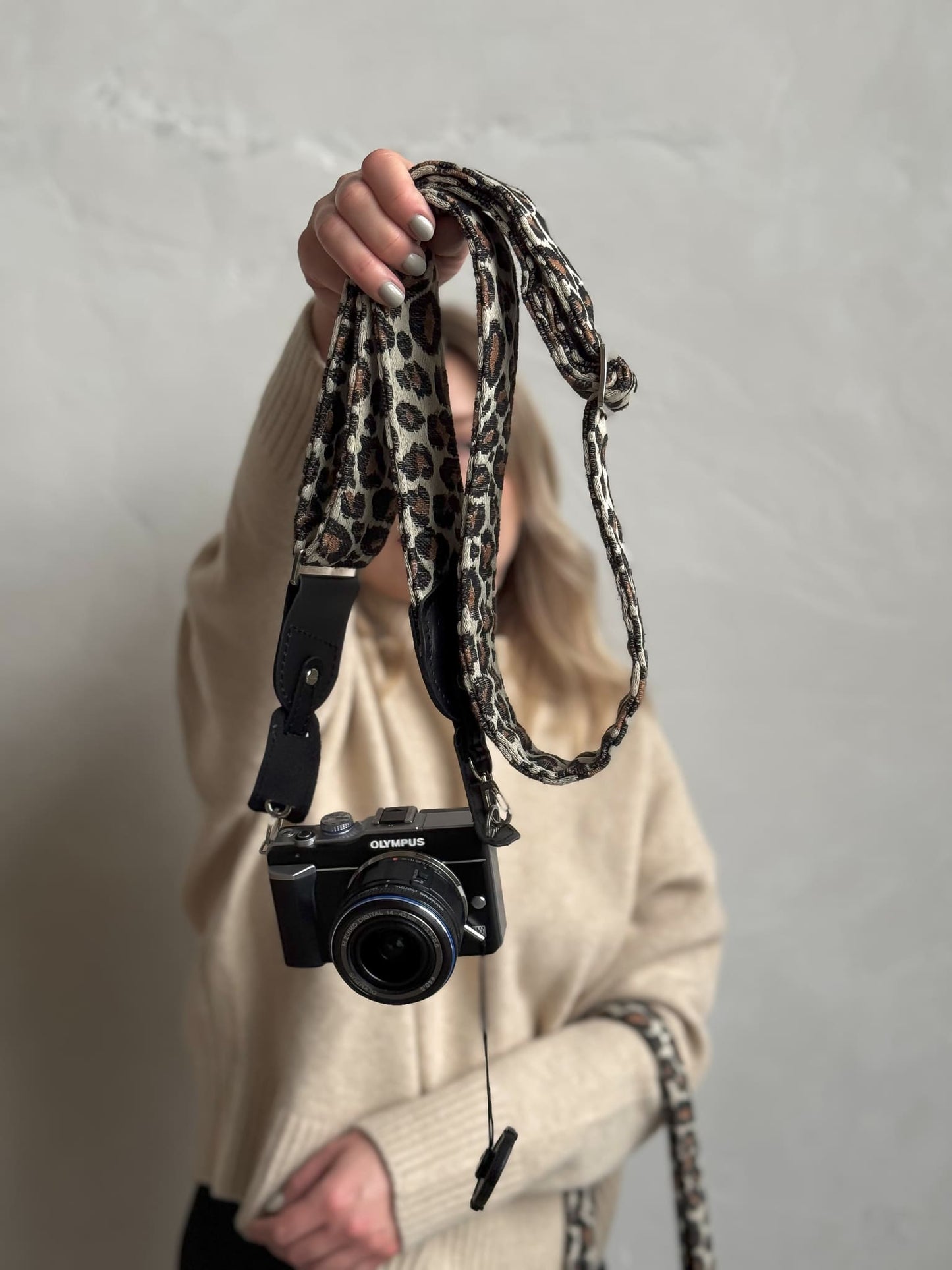 A person holds up a black mirrorless camera with a leopard-print camera strap against a light gray textured wall.