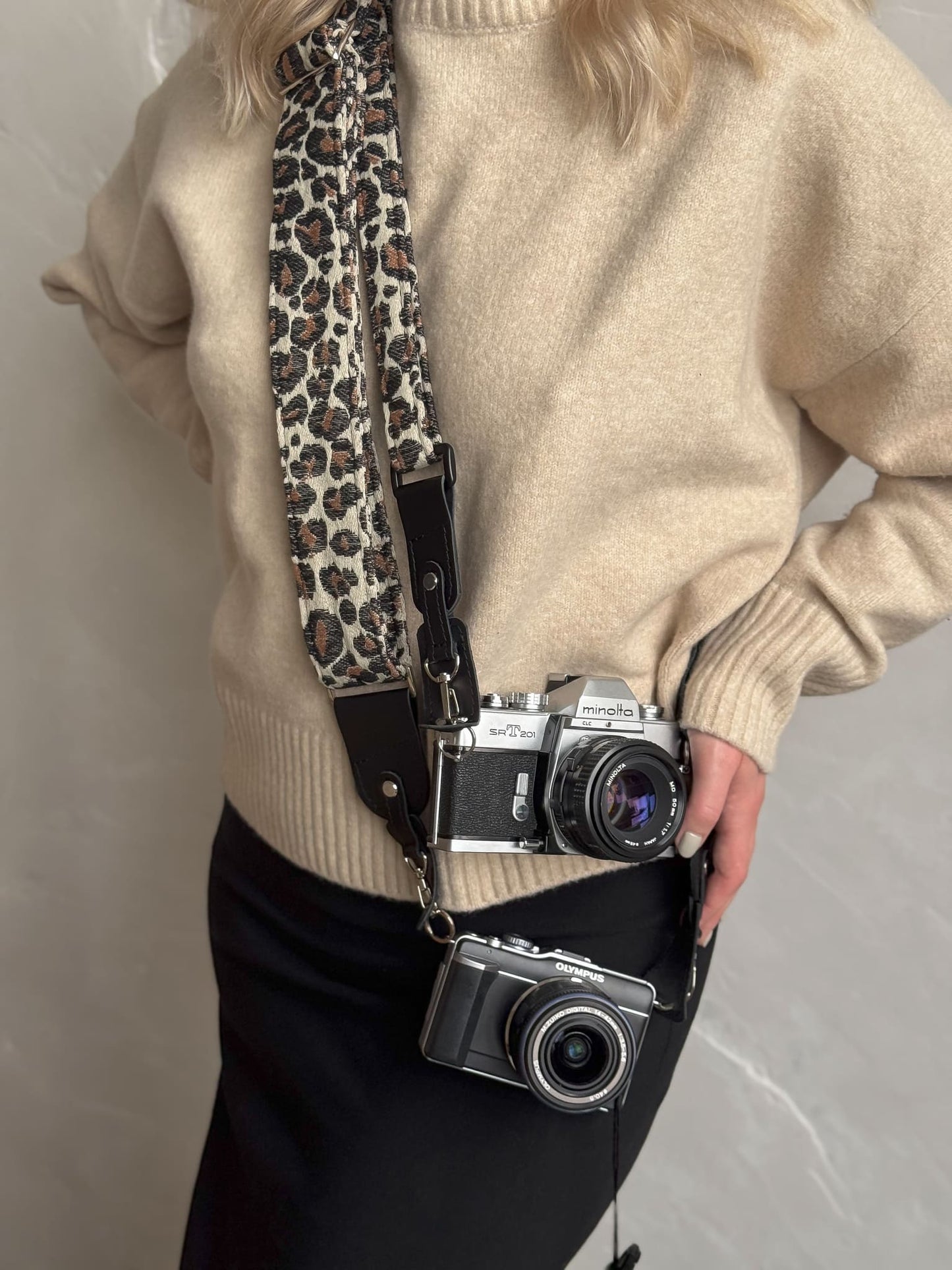 A person carries two cameras on a leopard-print camera strap. The strap features a beige base with brown, black, and white leopard spots, with black leather ends and hardware.