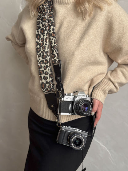 A person carries two cameras on a leopard-print camera strap. The strap features a beige base with brown, black, and white leopard spots, with black leather ends and hardware.