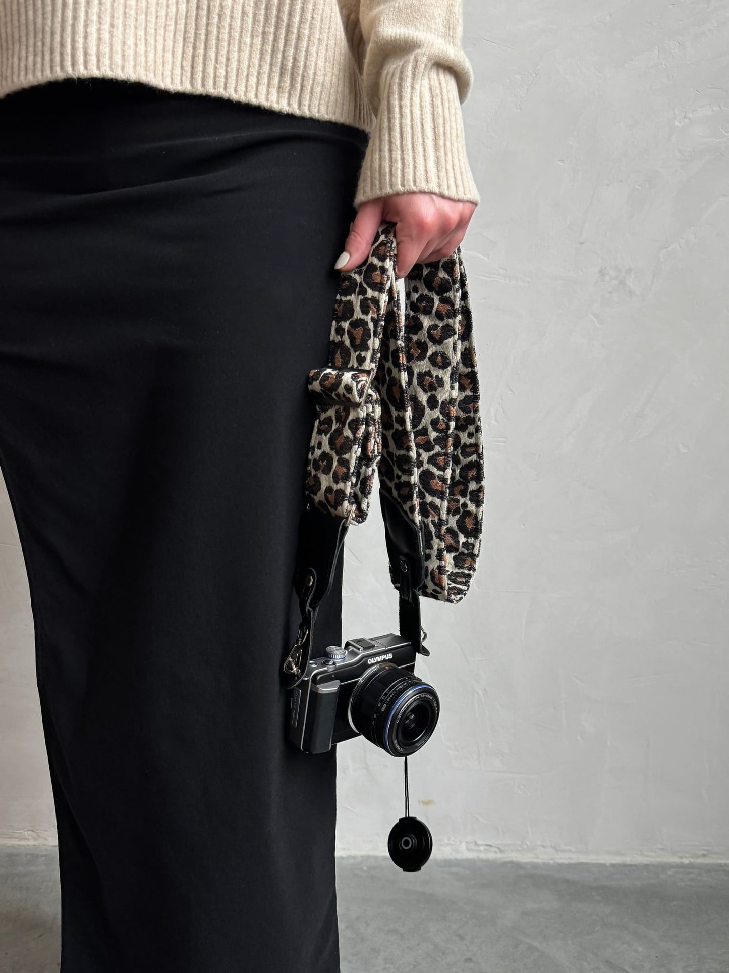A person holds a black camera with a leopard-print fabric strap against a light gray wall.