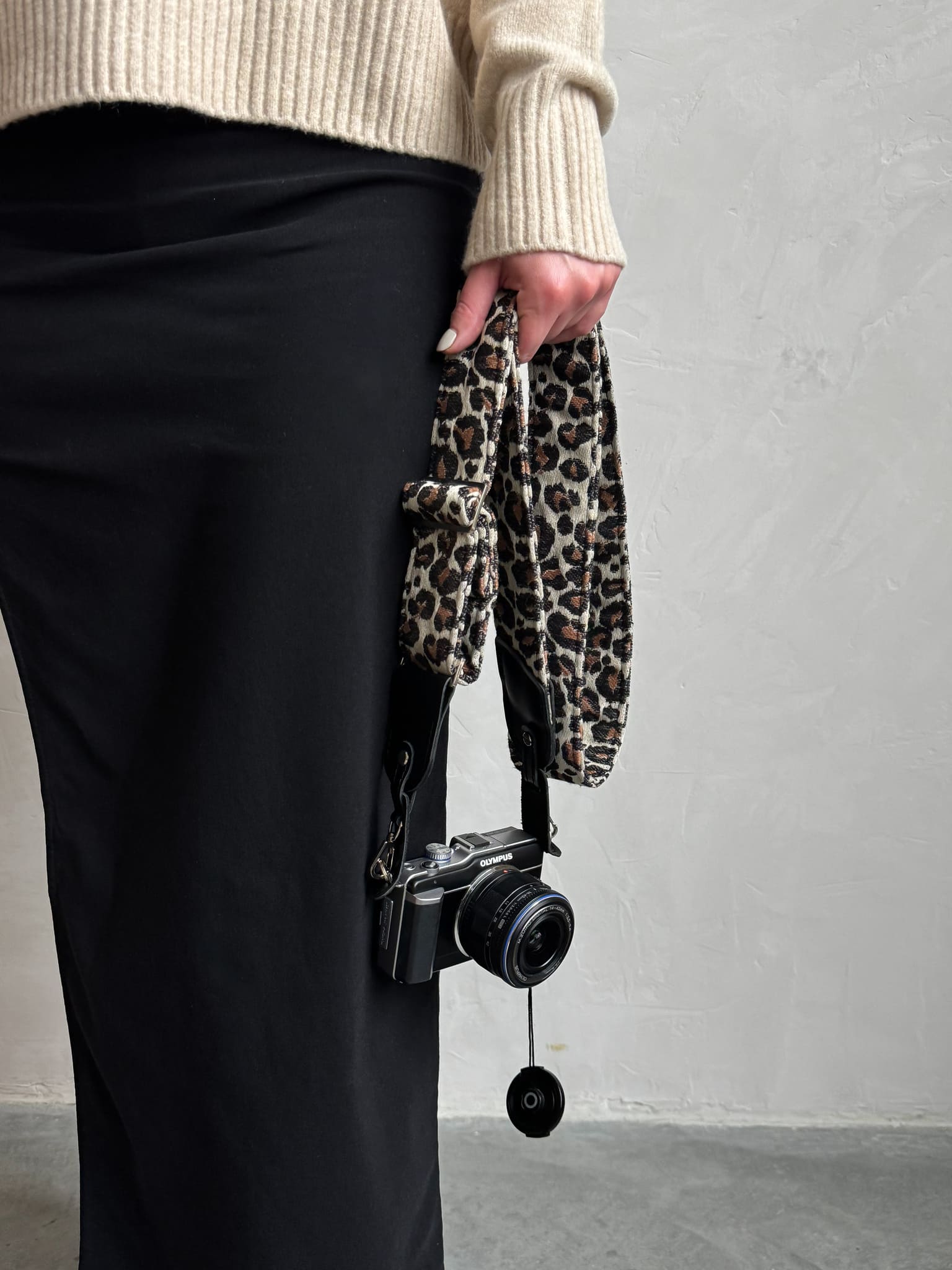 A person holds a black camera with a leopard-print fabric strap against a light gray wall.