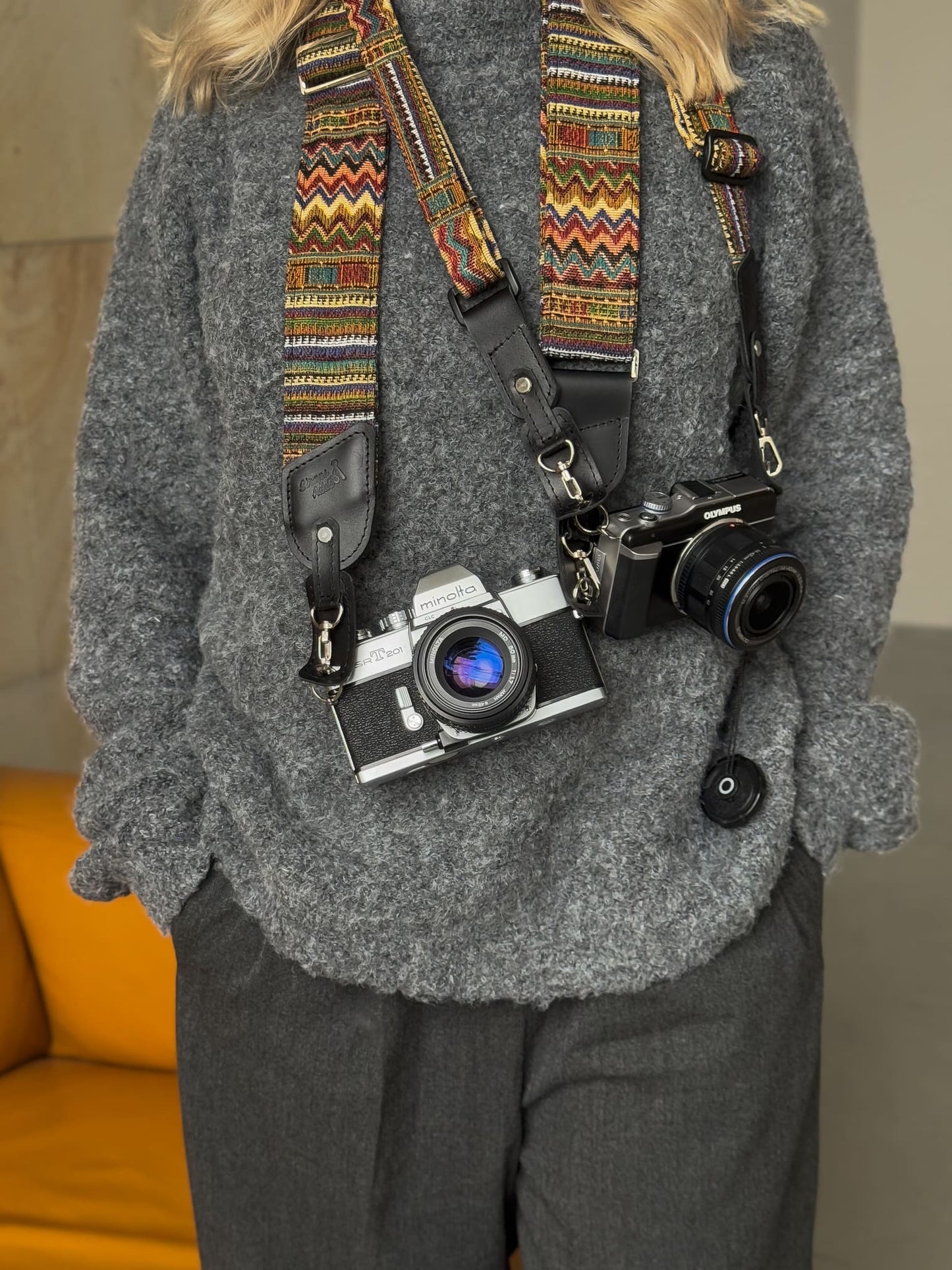 A person with two vintage-style cameras hanging from a colorful patterned camera straps across their chest. A mustard yellow chair is visible in the background.