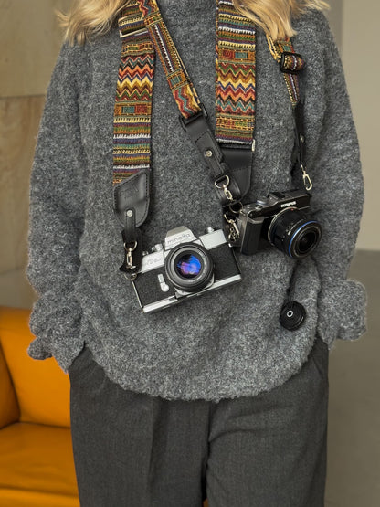 A person with two vintage-style cameras hanging from a colorful patterned camera straps across their chest. A mustard yellow chair is visible in the background.