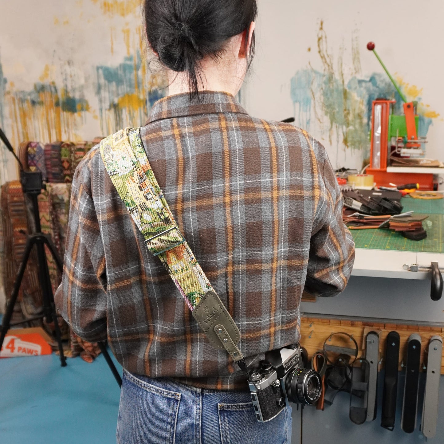 A person viewed from behind wearing a brown and gray plaid flannel shirt and blue jeans, carrying a camera over their shoulder with a decorative canvas-printed camera strap. The background shows a creative studio or workshop environment with a workbench, tools, and abstract wall art.