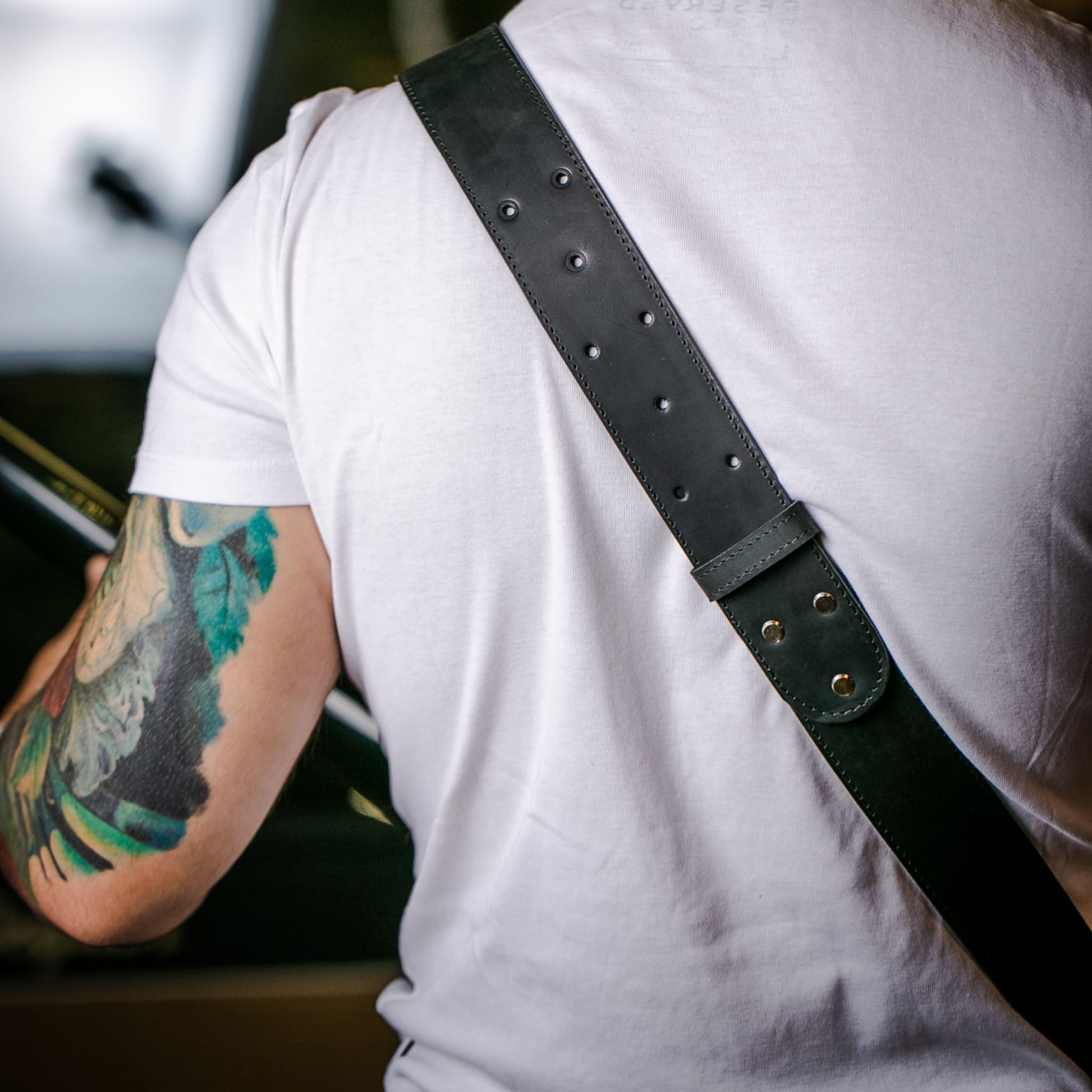 A close-up lifestyle shot of a person showing a black leather guitar strap with metal studs and rivets worn across their back.