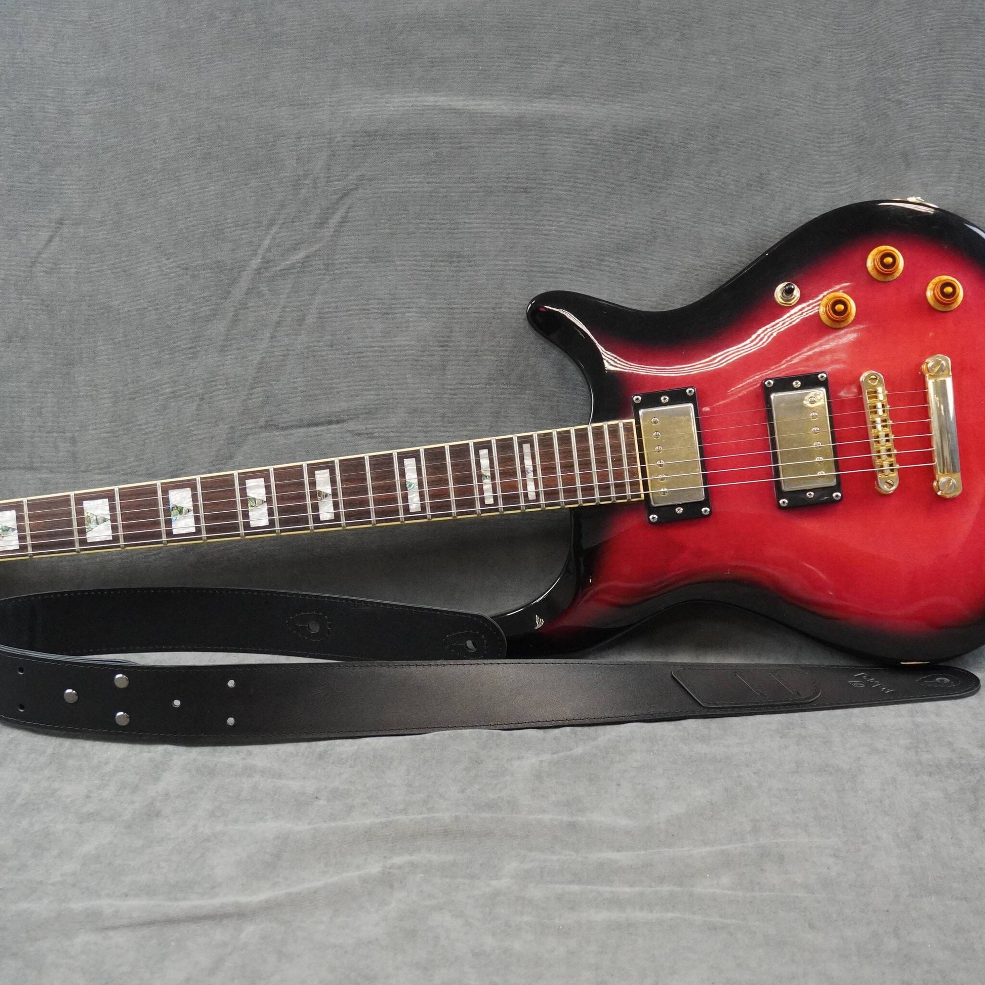 A red electric guitar with a black body finish and a black leather guitar strap, photographed on a gray fabric background.