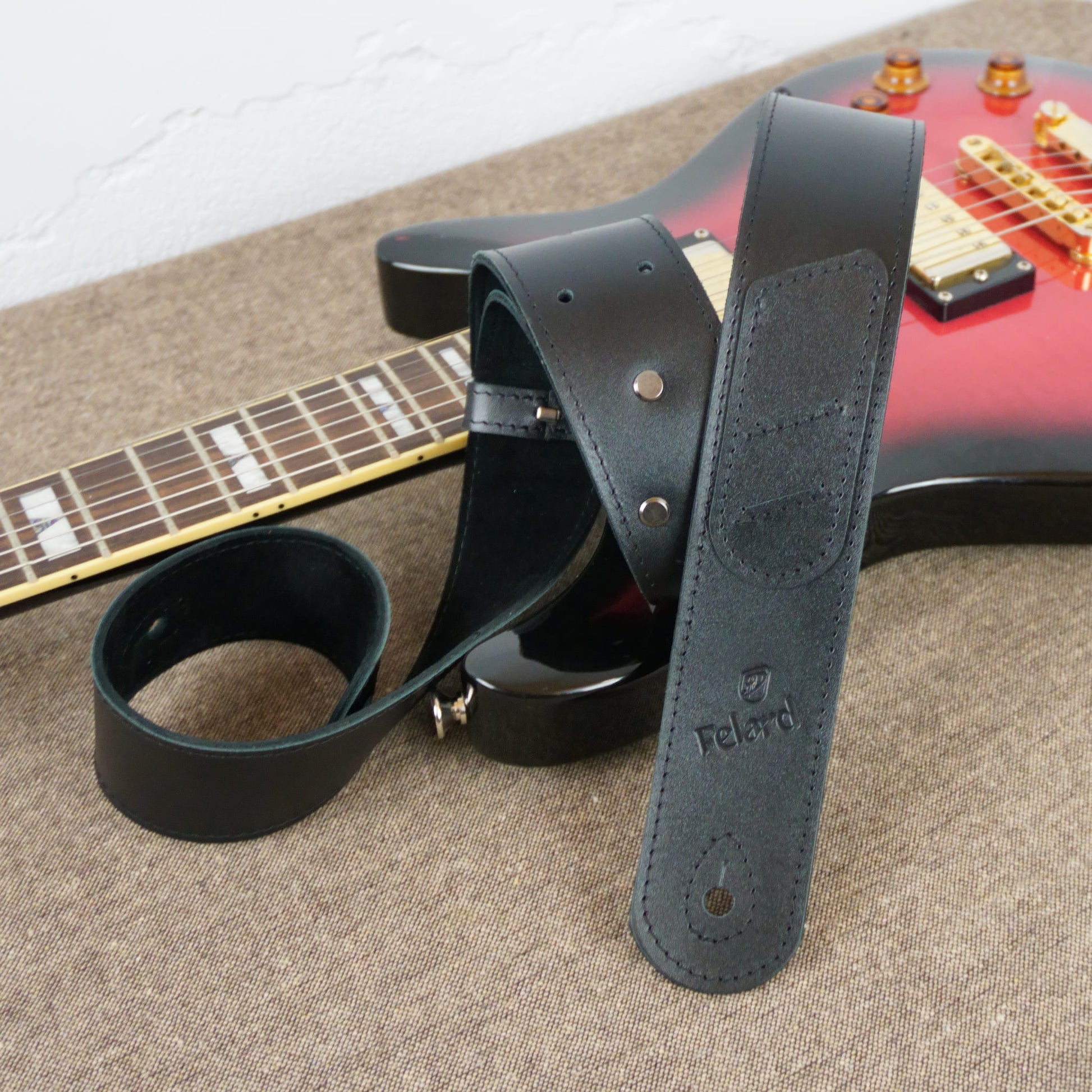 A black leather guitar strap with metal rivets and an embossed 'Felard' logo, draped over a red electric guitar on a textured beige fabric surface.