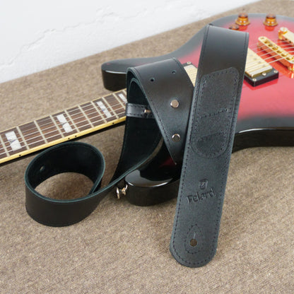 A black leather guitar strap with metal rivets and an embossed 'Felard' logo, draped over a red electric guitar on a textured beige fabric surface.